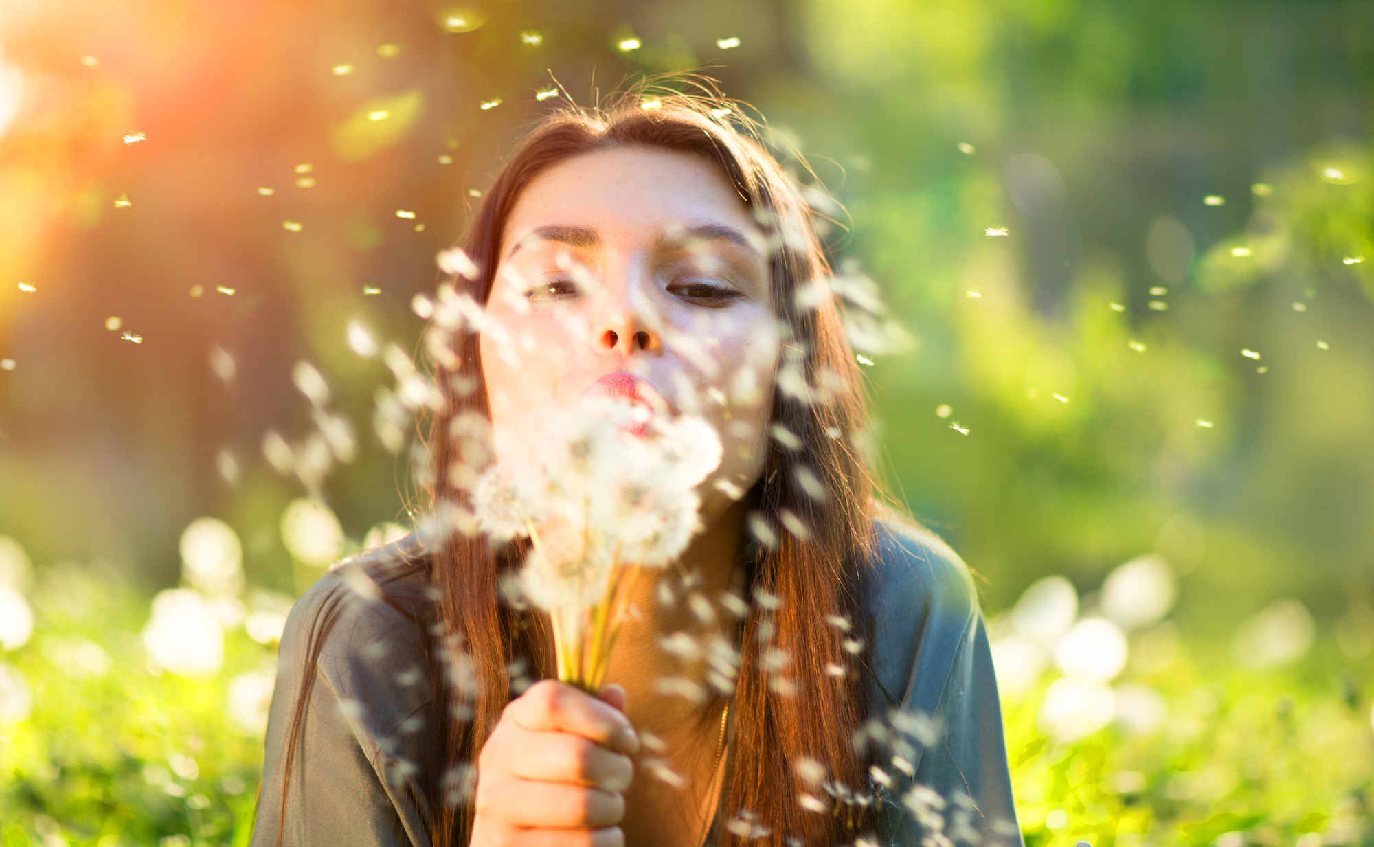 Eine junge Frau mit langen, braunen Haaren bläst in einem sonnigen Feld Pusteblumen, deren Samen durch die Luft wirbeln. Sie symbolisiert einen allergiefreien Start in Frühling und Sommer.