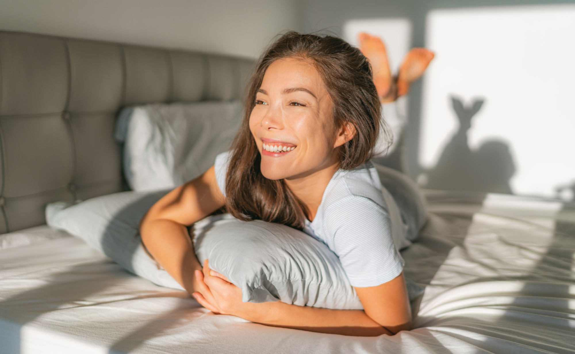 Lächelnde Frau liegt entspannt in einem sonnigen Schlafzimmer auf dem Bauch im Bett, den Kopf auf ein Kissen gestützt. Sie symbolisiert Ausgeruhtheit durch gute Schlafhygiene,