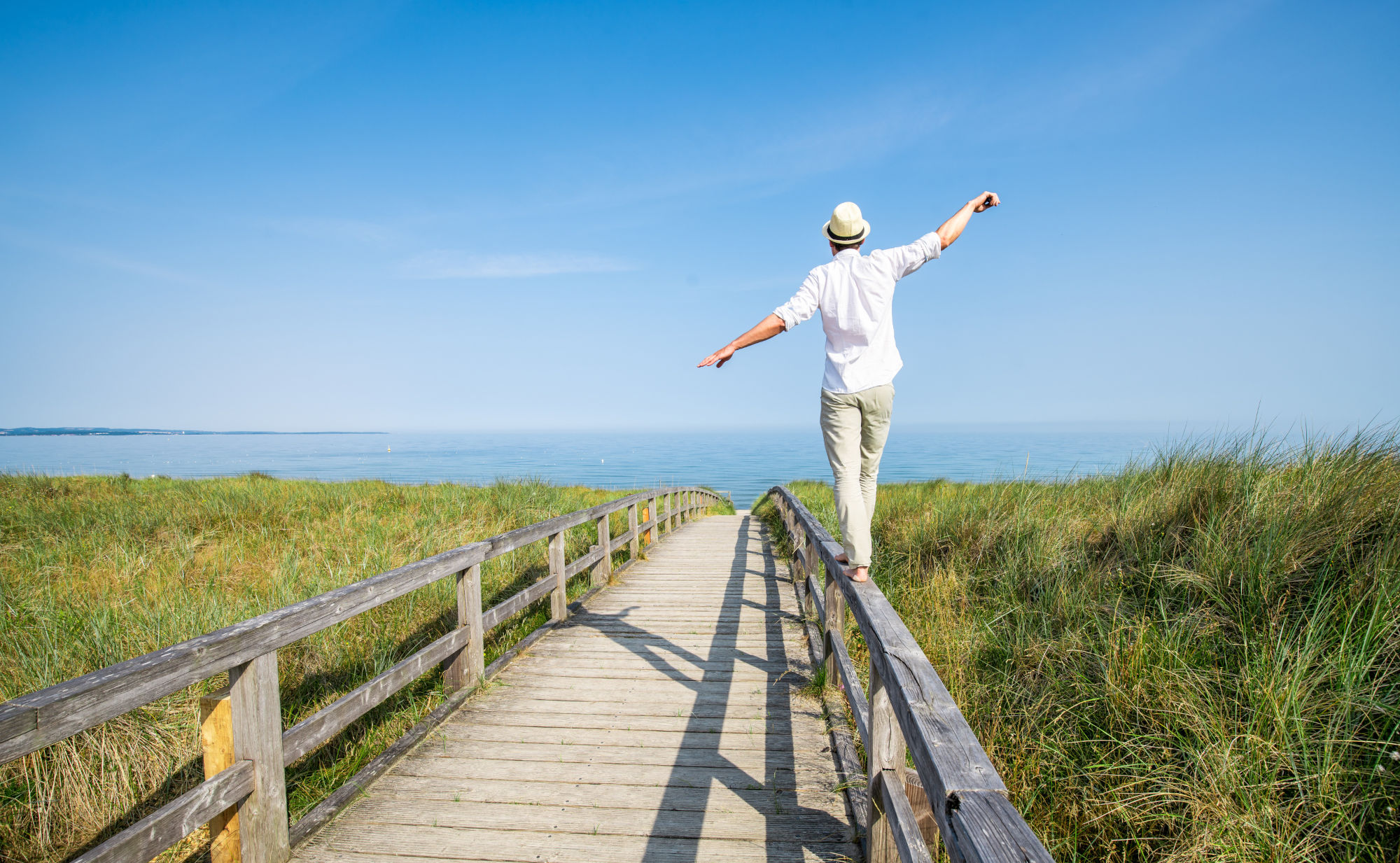 Ein Mensch mit hellem Hemd und Hut balanciert mit ausgebreiteten Armen auf dem Geländer eines Holzstegs, der durch Dünen zum Meer führt. Symbolfoto für die Basen-Balance im Körper.