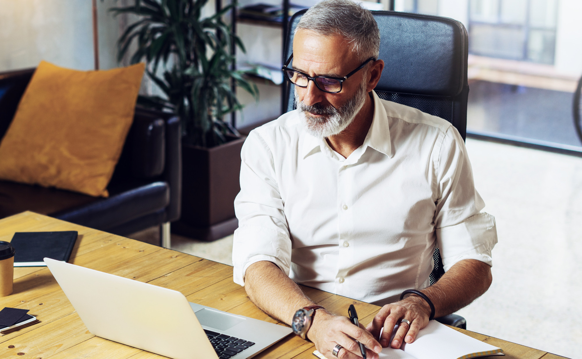 Ein Mann mit Brille sitzt an einem Holztisch, schaut auf einen Laptop und macht sich Notizen in ein Notizbuch. Ein Symbolfoto für Gesundheit durch Wissen über die eigenen Cholesterinwerte.