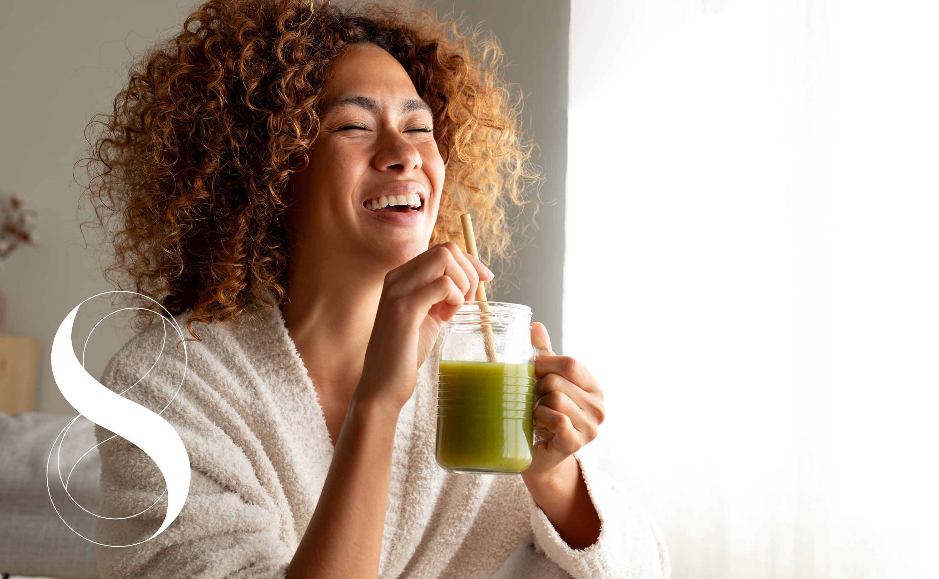 Eine lachende Frau im Bademantel führt ein Glas zum Mund, das mit frischem, grünem Smoothie gefüllt ist.