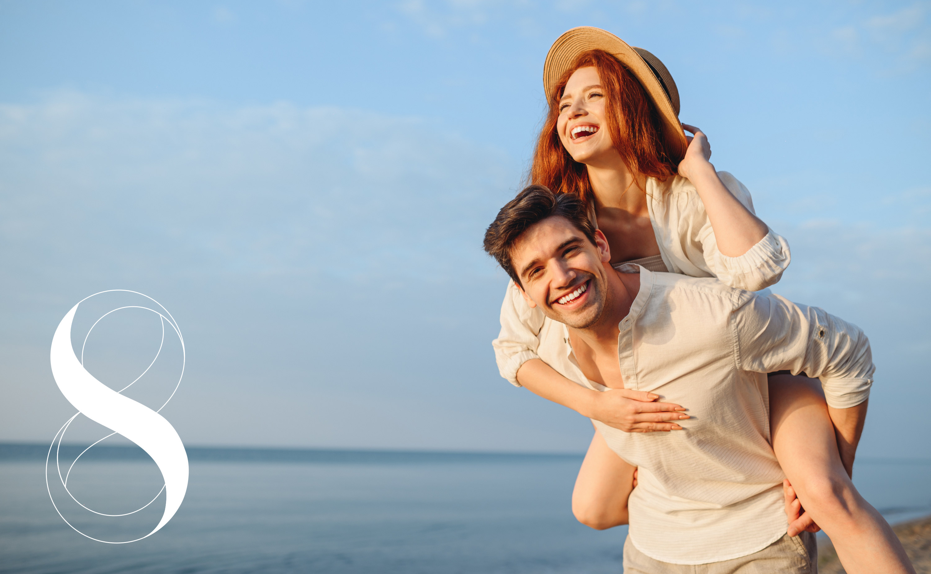 Ein lächelnder Mann trägt eine lachende Frau mit roten Haaren und Strohhut Huckepack am Strand, das Meer im Hintergrund. Ein Symbolfoto von LIFE LIGHT für gesundes Gewicht vor dem und während des Urlaubs.
