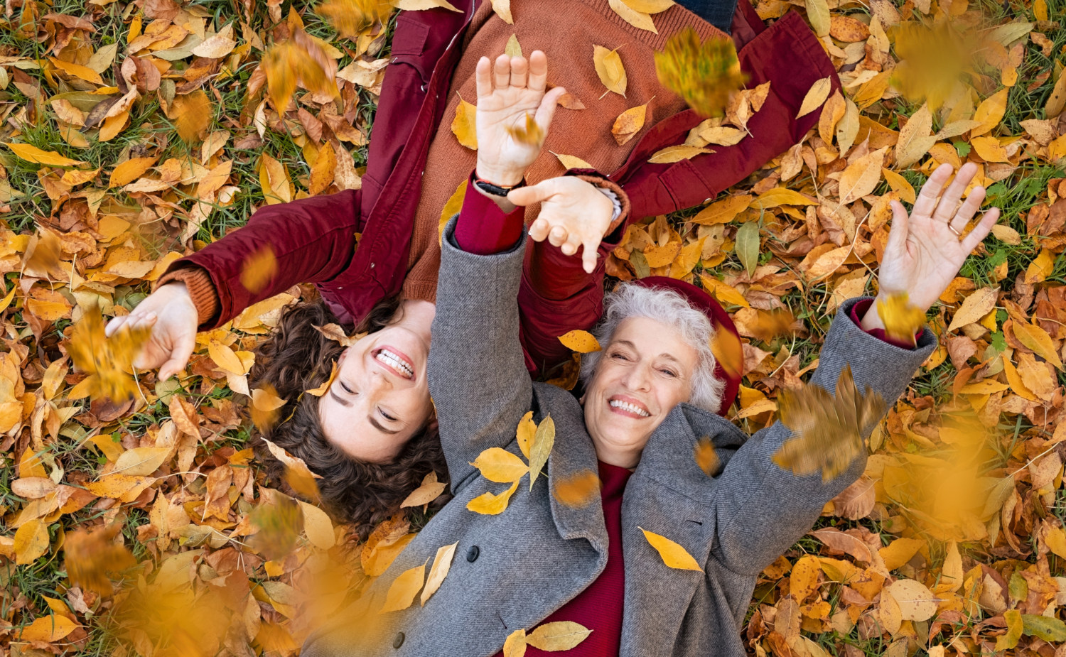 Zwei lächelnde Frauen liegen auf dem Rücken auf einem Teppich aus herbstlichen Blättern, sie werfen Blätter in die Luft. Symbolfoto für starke Abwehrkräfte im Herbst.