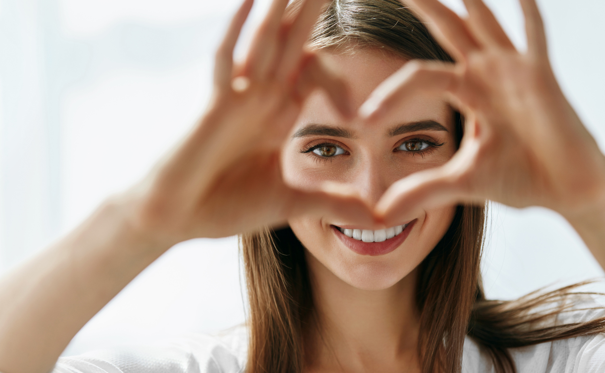 Eine junge Frau lächelt und formt mit ihren Händen ein Herz vor ihrem Gesicht, durch das man ihre Augen sieht. Sie symbolisiert gesunde Augen und klare Sicht durch entsprechende Mikronährstoffe.