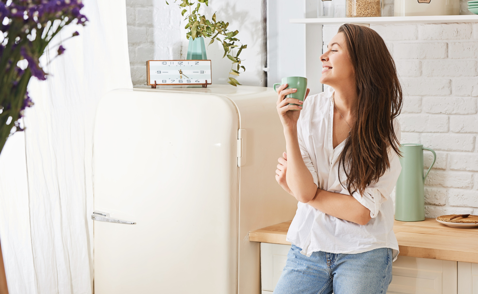 Eine junge Frau mit langen, braunen Haaren steht in einer Küche, lächelt und hält eine Teetasse in der Hand. Ein Symbolfoto von LIFE LIGHT für Wohlbefinden mit einer gesunden Harnblase.