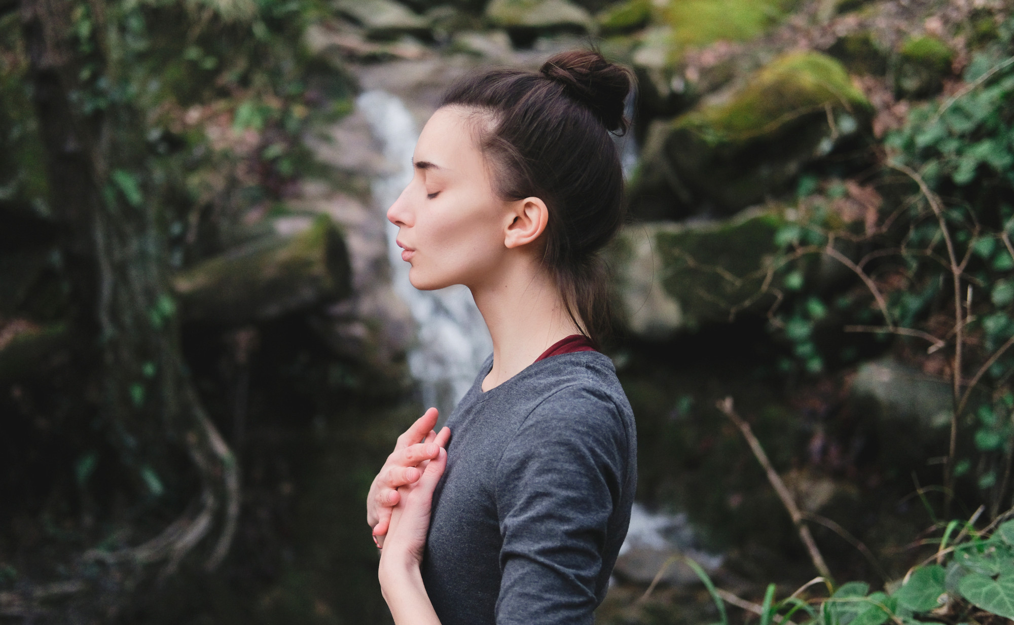 Eine Frau steht mit geschlossenen Augen und gefalteten Händen vor der Brust in der Natur, im Hintergrund ist ein kleiner Wasserfall. Ein Symbolfoto von LIFE LIGHT für eine gesunde Lunge und frische Luft.