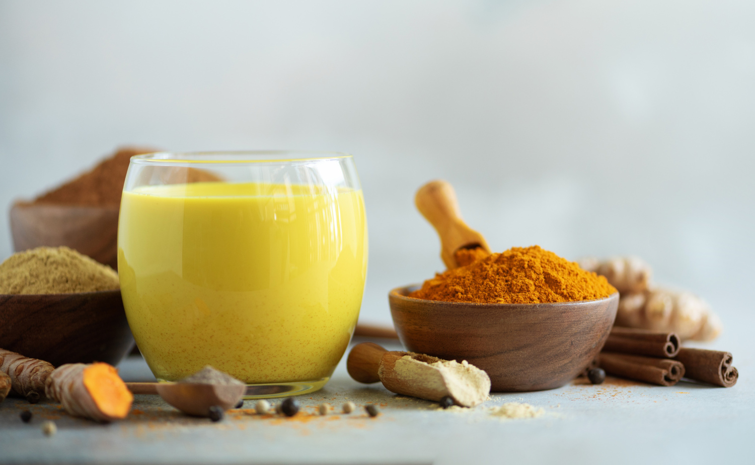 Ein Glas goldene Milch, daneben Macapulver und Kurkumapulver in 2 Schalen, Zimtstangen und Kurkuma-Wurzelknollen im Vordergrund, der Hintergrund ist hellblau.