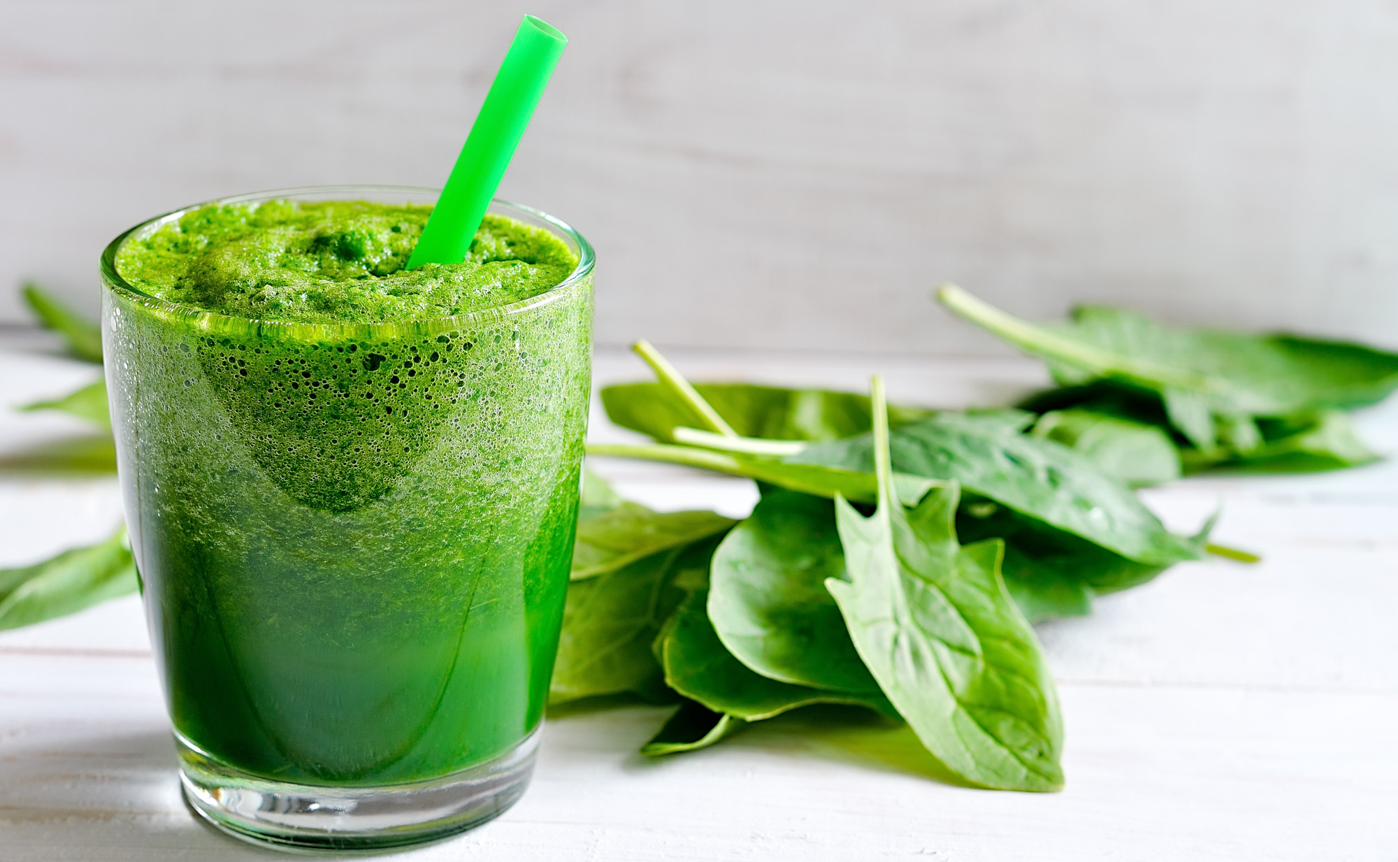 Ein Glas mit einem leuchtend grünen Smoothie und einem grünen Strohhalm, umgeben von frischen Spinatblättern. Symbolfoto für grüne, gesunde Superfoods zum Trinken.