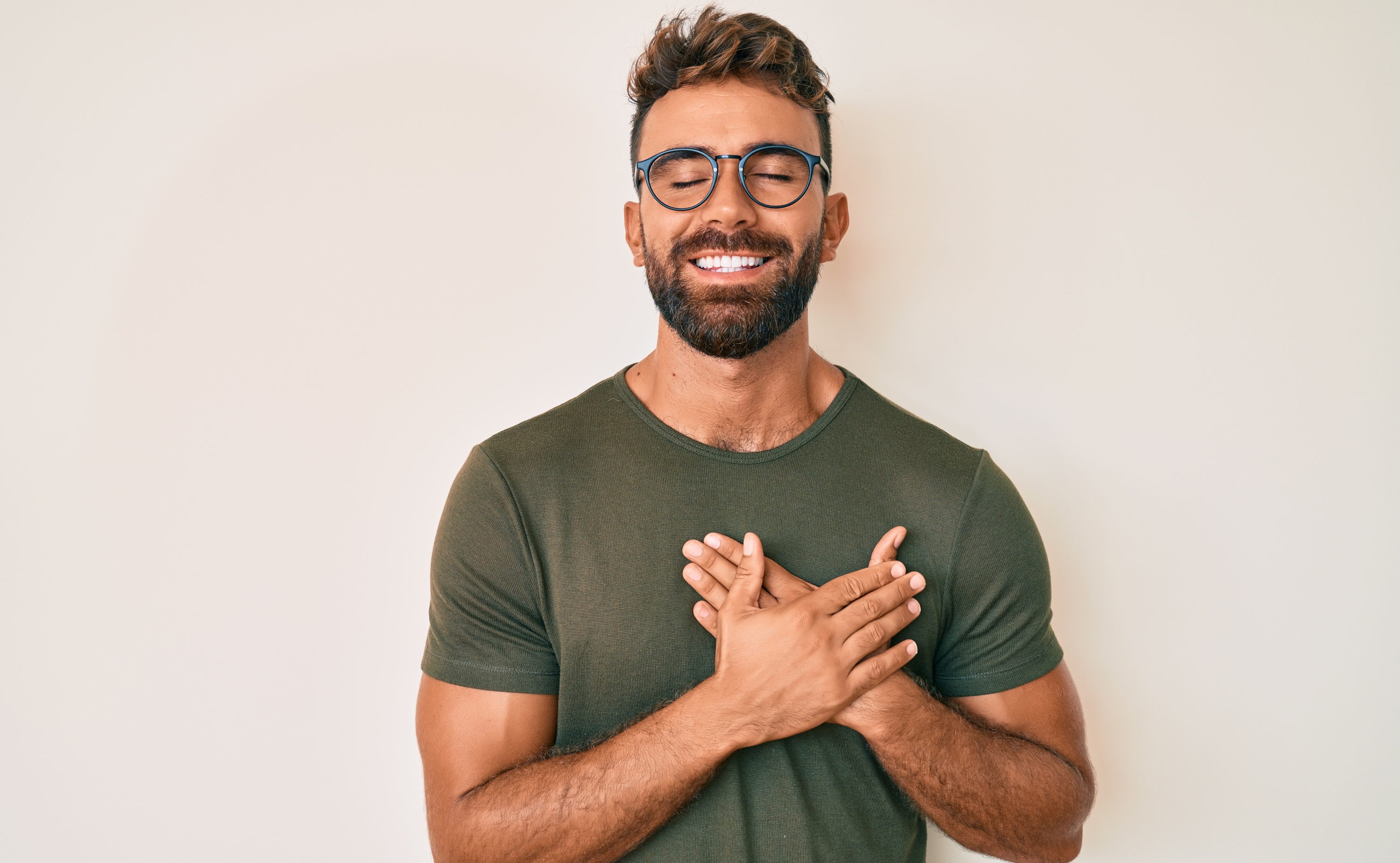 Ein lächelnder Mann mit Brille und Bart vor hellem Hintergrund hält mit geschlossenen Augen beide Hände über seinem Herzen. Ein Symbolfoto für ein gesundes Herz in der Brust.