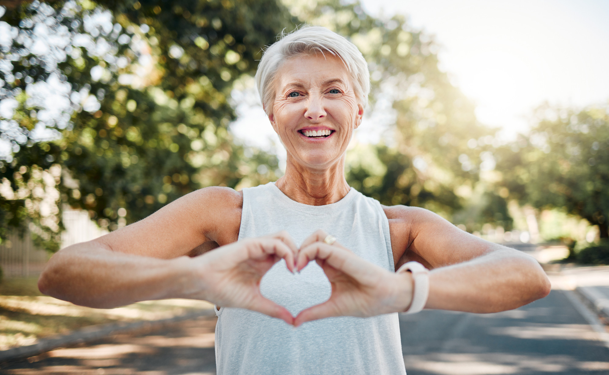 Eine lächelnde Frau mit kurzen, grauen Haaren in Sportkleidung, ihre Hände formen ein Herz. Im Hintergrund sind Bäume und die strahlende Sonne. Ein Symbolfoto von LIFE LIGHT zur Herzgesundheit an heißen Tagen.