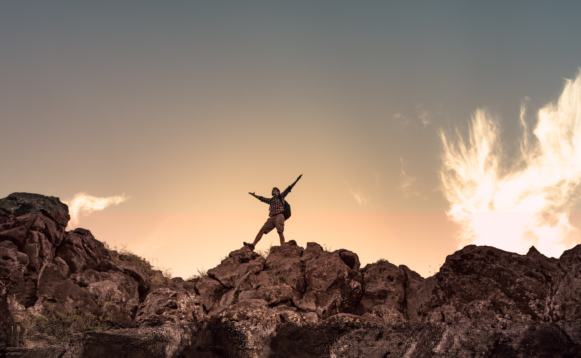 Ein Wanderer steht auf einem felsigen Berggipfel und breitet die Arme aus, während die Sonne am Horizont untergeht. Ein Symbolfoto für die Regeneration der menschlichen Zellktaftwerke.