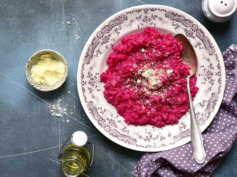 Ein Teller mit leuchtend rotem Rote-Bete-Risotto, bestreut mit Käse, steht neben einer Schale mit geriebenem Käse und einer Flasche Öl.